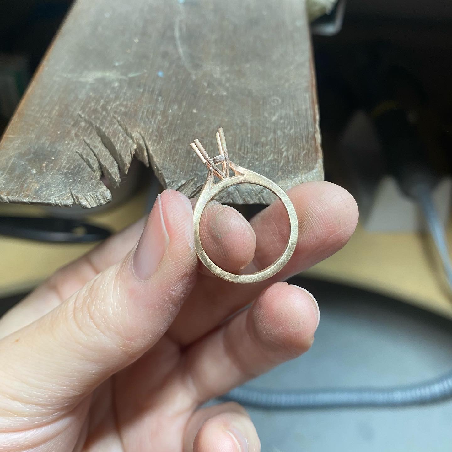 A hand holds an unfinished ring — a plain band with an empty four-claw setting where the stone will go — in front of a worn wooden bench peg.