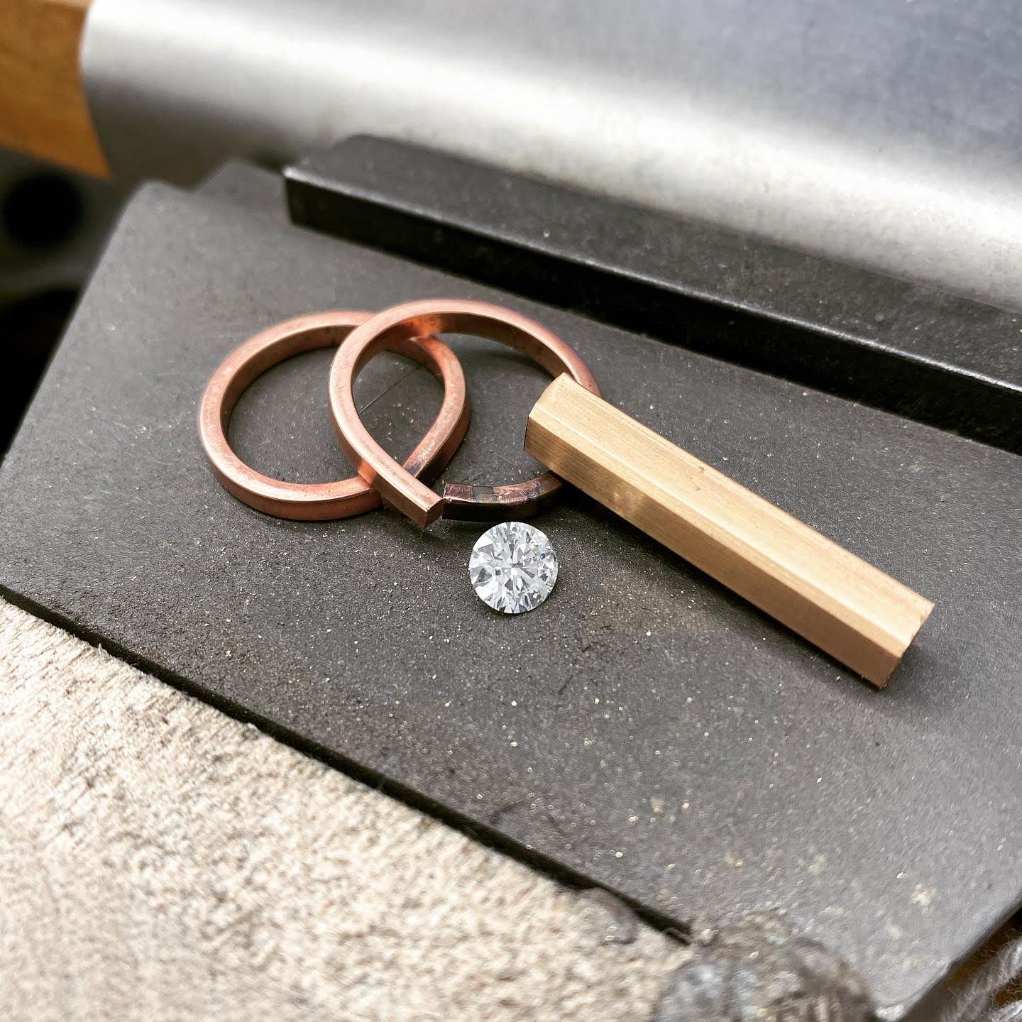 Two rose gold ring blanks, a loose round brilliant diamond and a rectangular gold ingot laid out together on a steel bench block.