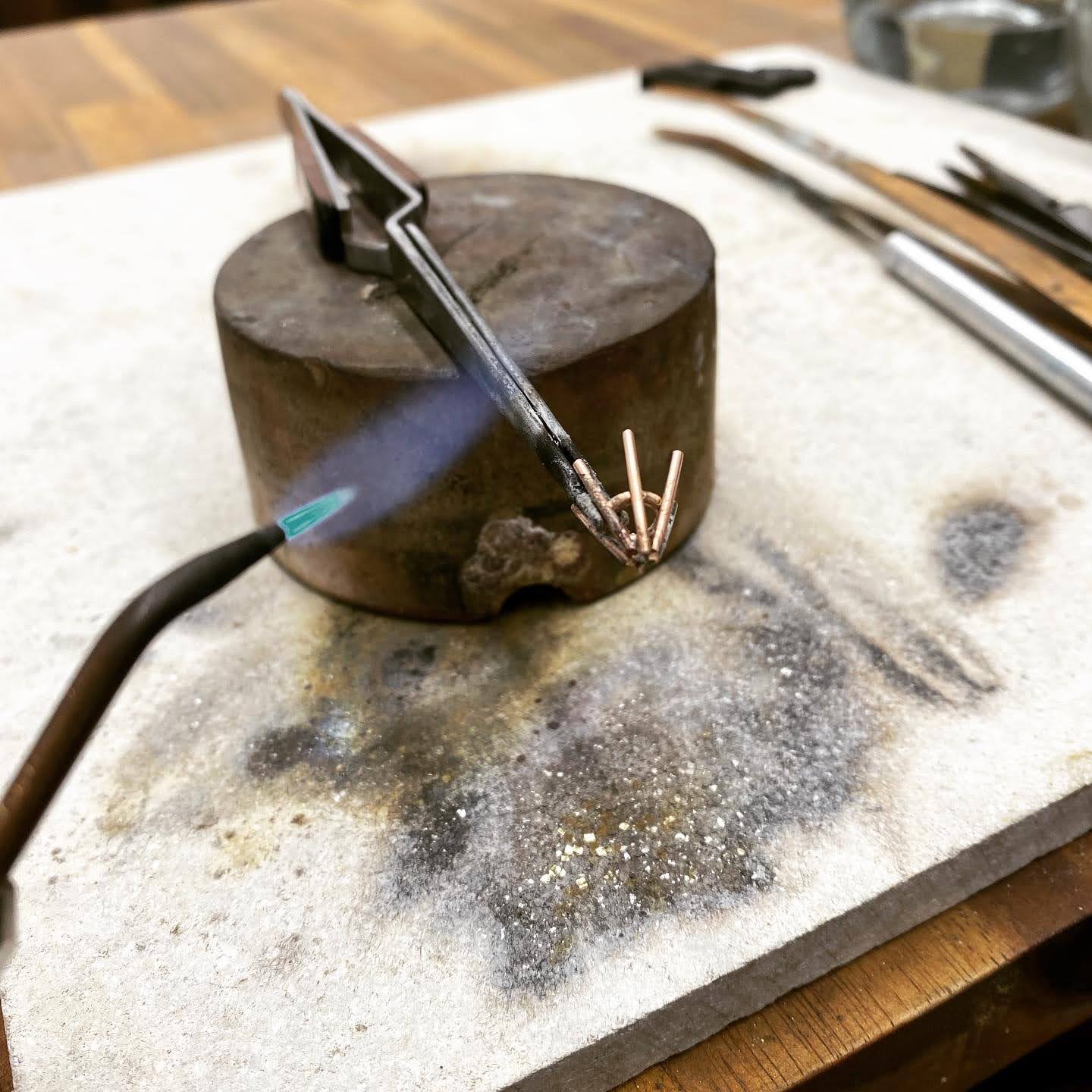 A jeweller's torch plays a blue flame over a set of rose gold claws held in soldering tweezers on a scorched soldering block, with files and tools around it.