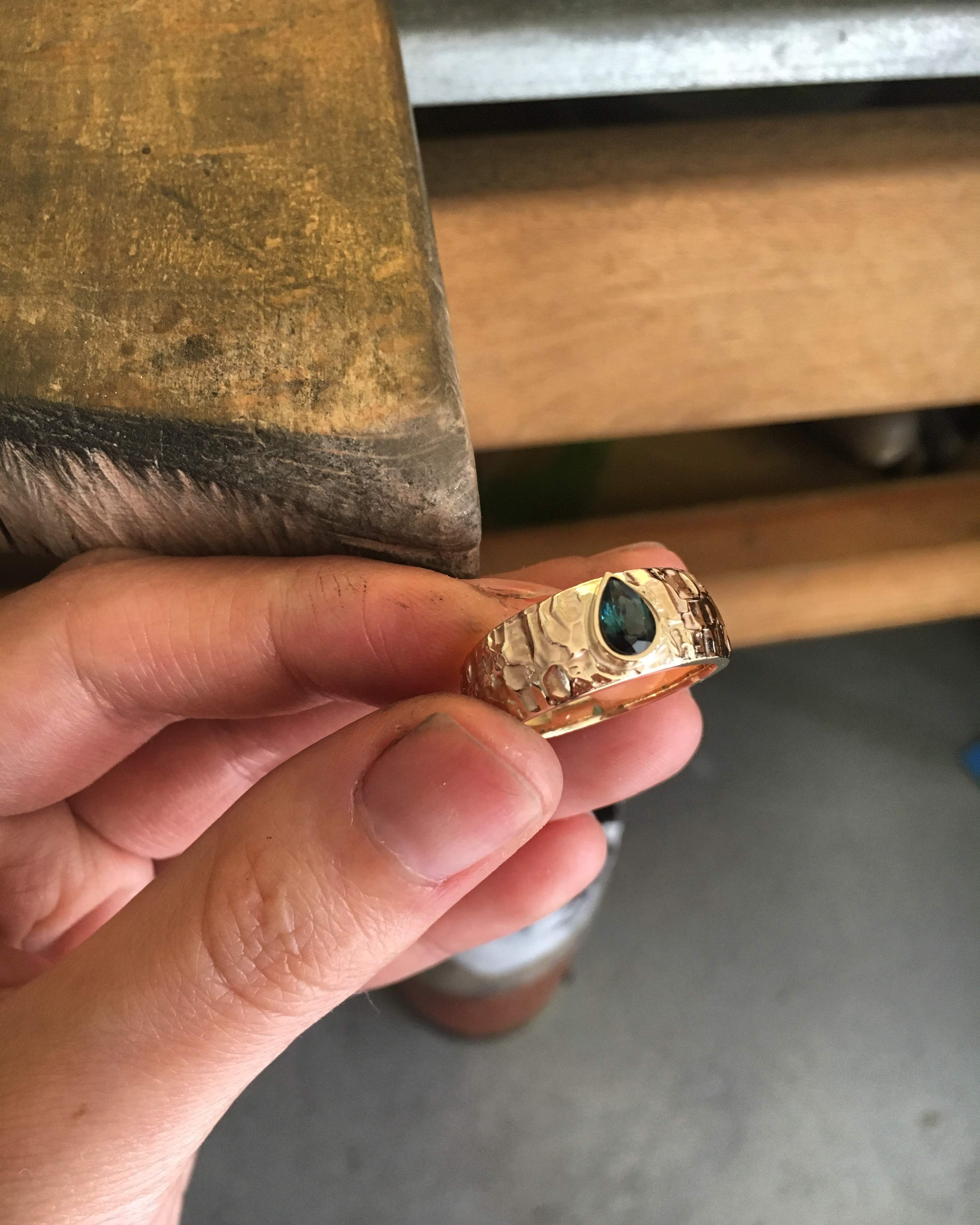 A hand holds a wide yellow gold ring with a hammered, bark-like texture, set with a pear-cut deep teal stone, in front of a jeweller's bench peg.