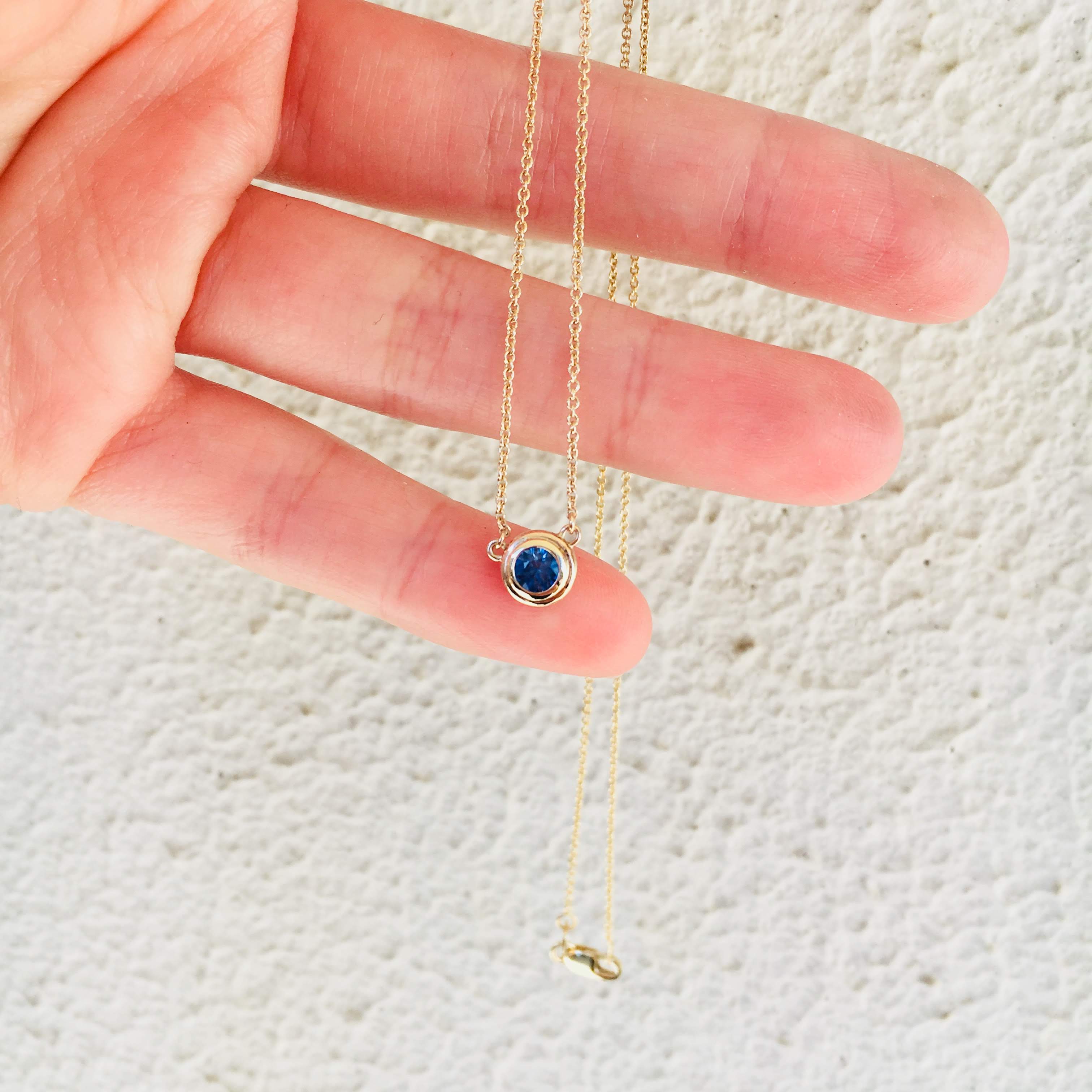 A fine gold chain held across an open palm, with a small round blue sapphire in a rubover setting hanging at its centre.
