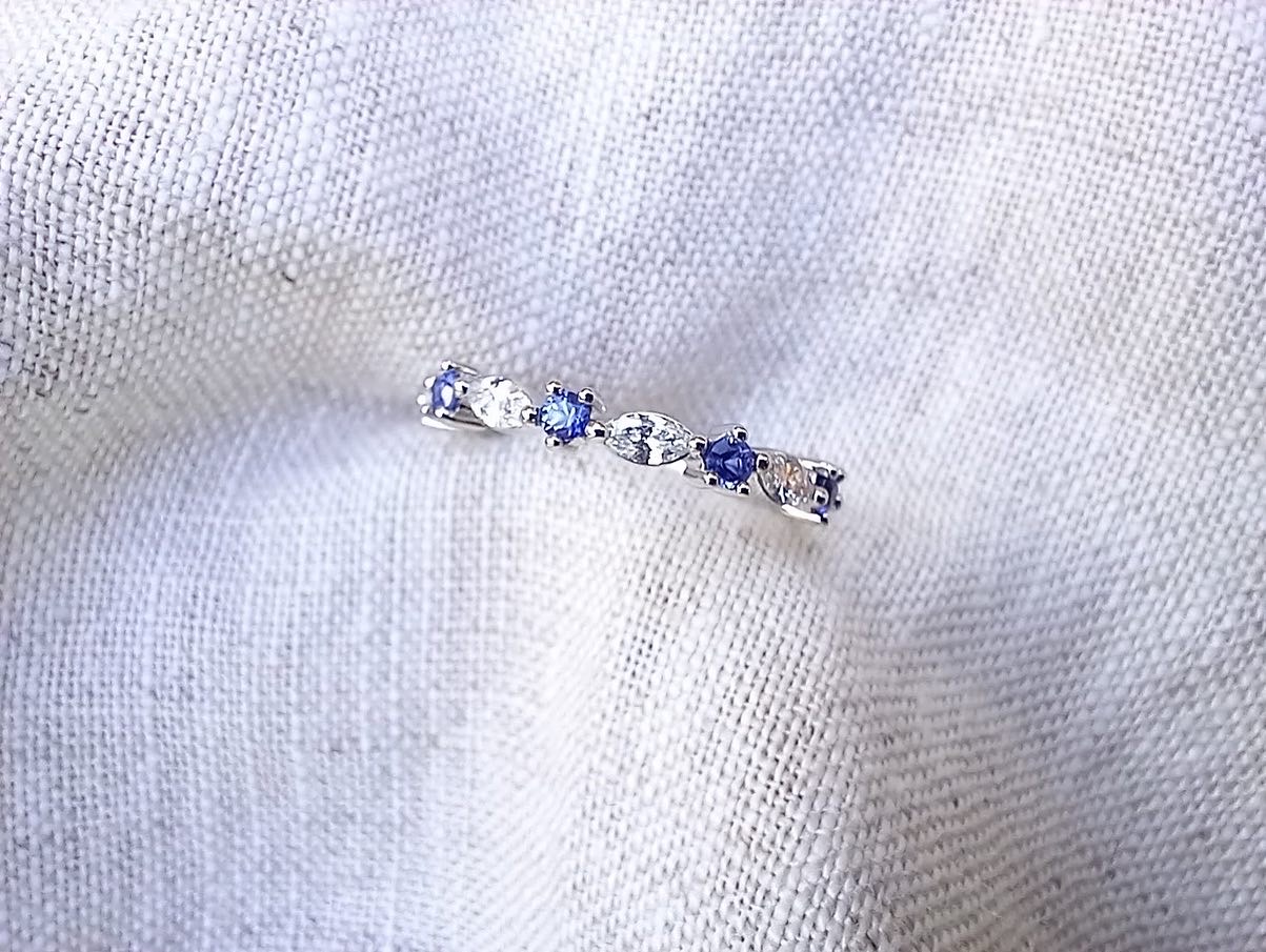 A slim white gold band set with an alternating row of blue sapphires and white marquise and round diamonds, lying on pale linen.