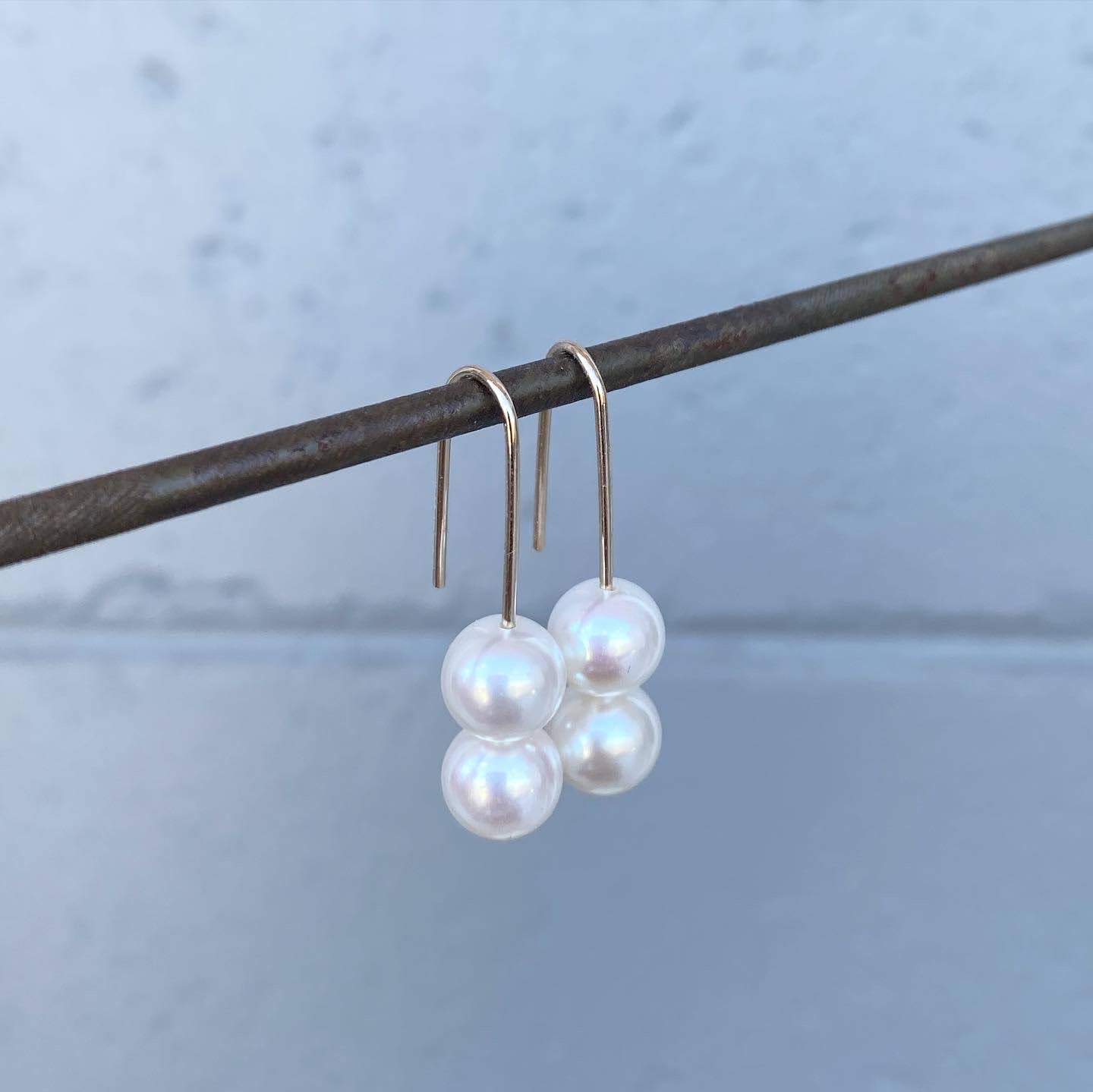 A pair of white pearl drop earrings on long gold hooks, hanging from a steel bar against a pale blue wall.