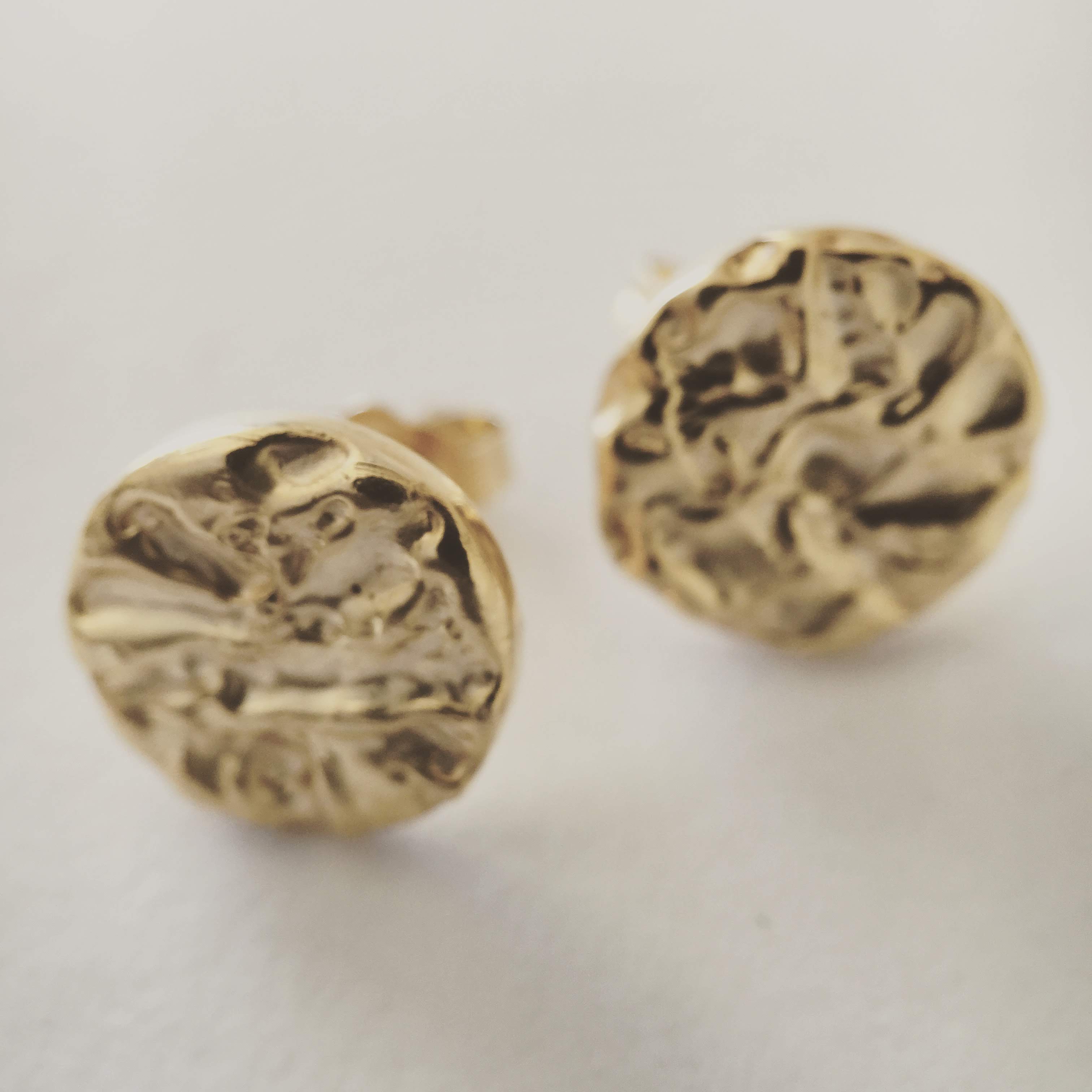 A pair of round yellow gold stud earrings with a heavily hammered, molten-looking surface that catches the light unevenly.