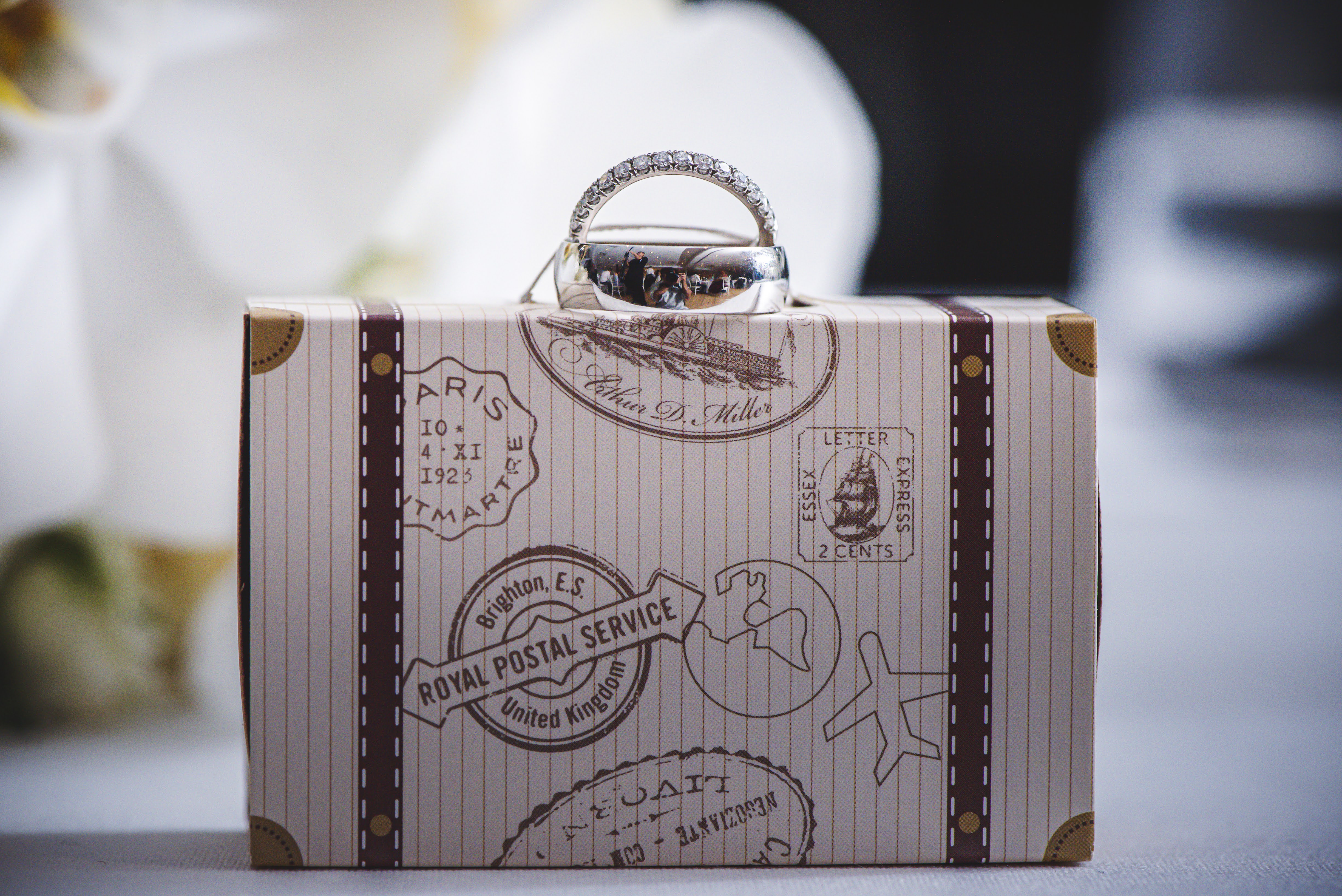 Two wedding bands, one set with a row of small diamonds and one plain, balanced on the handle of a miniature vintage suitcase printed with postal stamps, with white flowers behind.