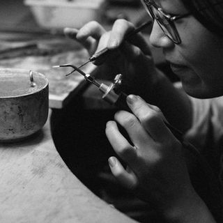 Black and white photograph of Lea at her bench in glasses, holding a ring in a clamp close to her face while she works on it with a fine tool.