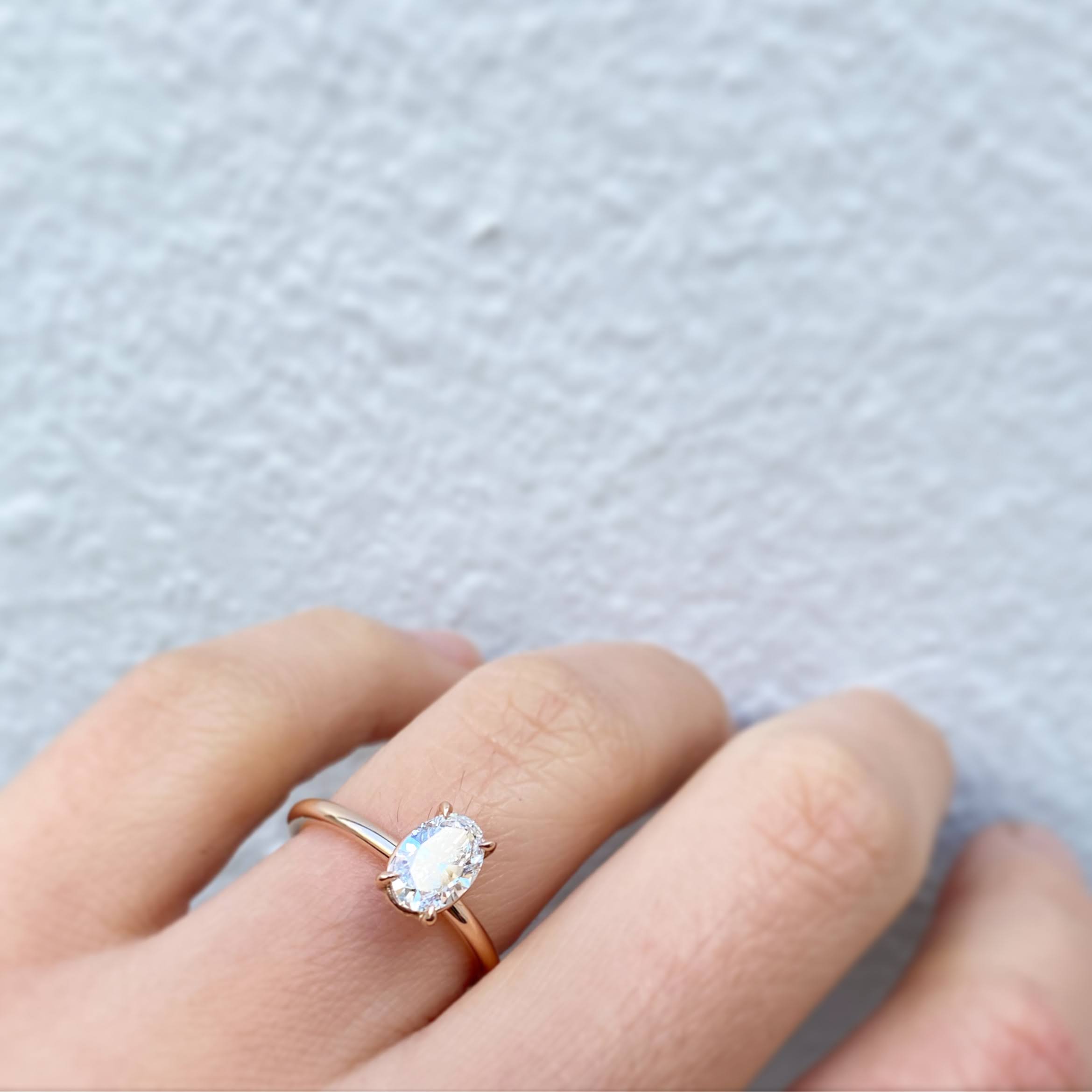 An oval brilliant-cut diamond in a claw setting on a slim yellow gold band, worn on a hand against a white rendered wall.