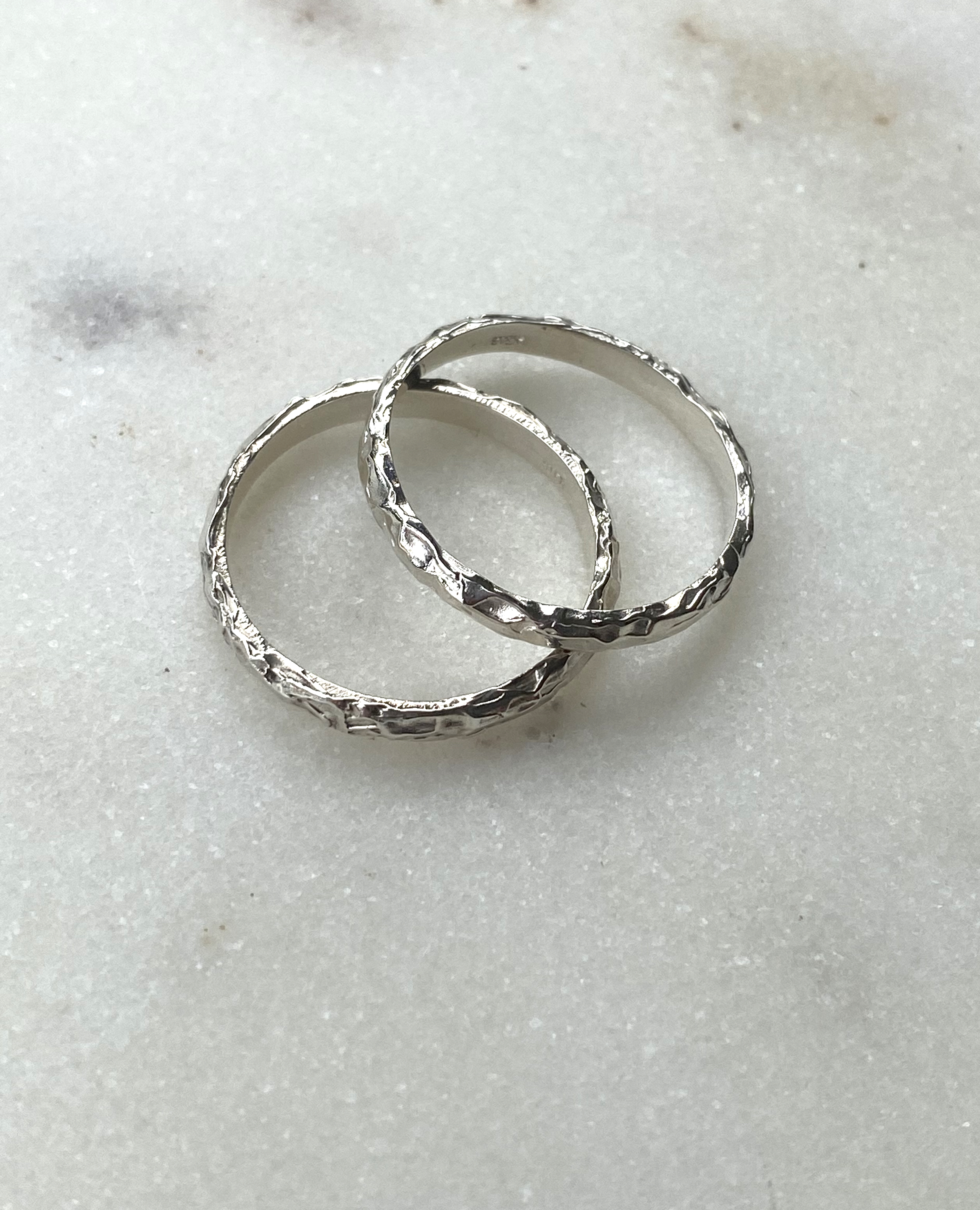Two white gold wedding bands with a rough, chiselled texture, resting together on white marble.