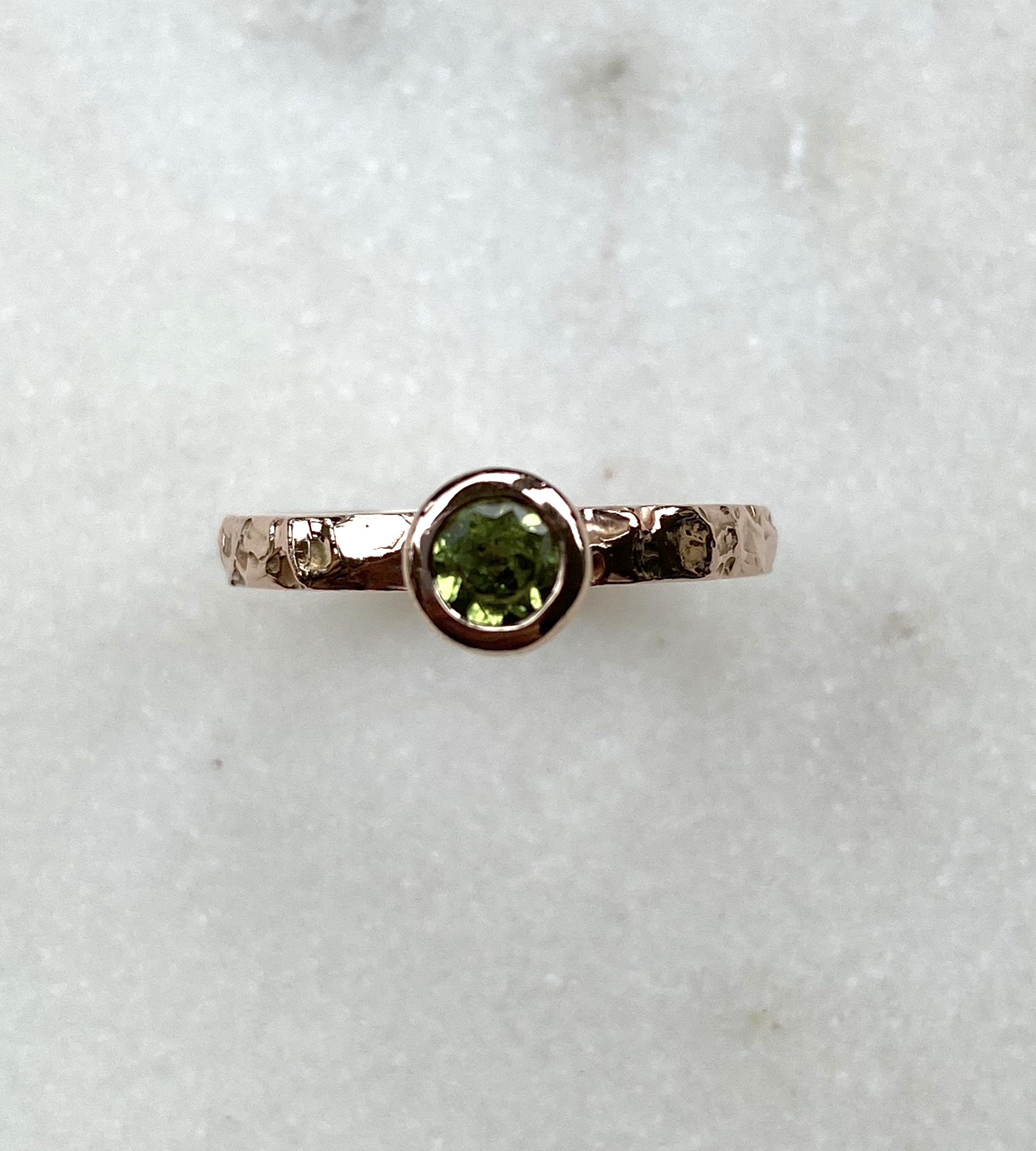 A round green tourmaline in a rose gold rubover setting on a heavily textured rose gold band, against a white background.