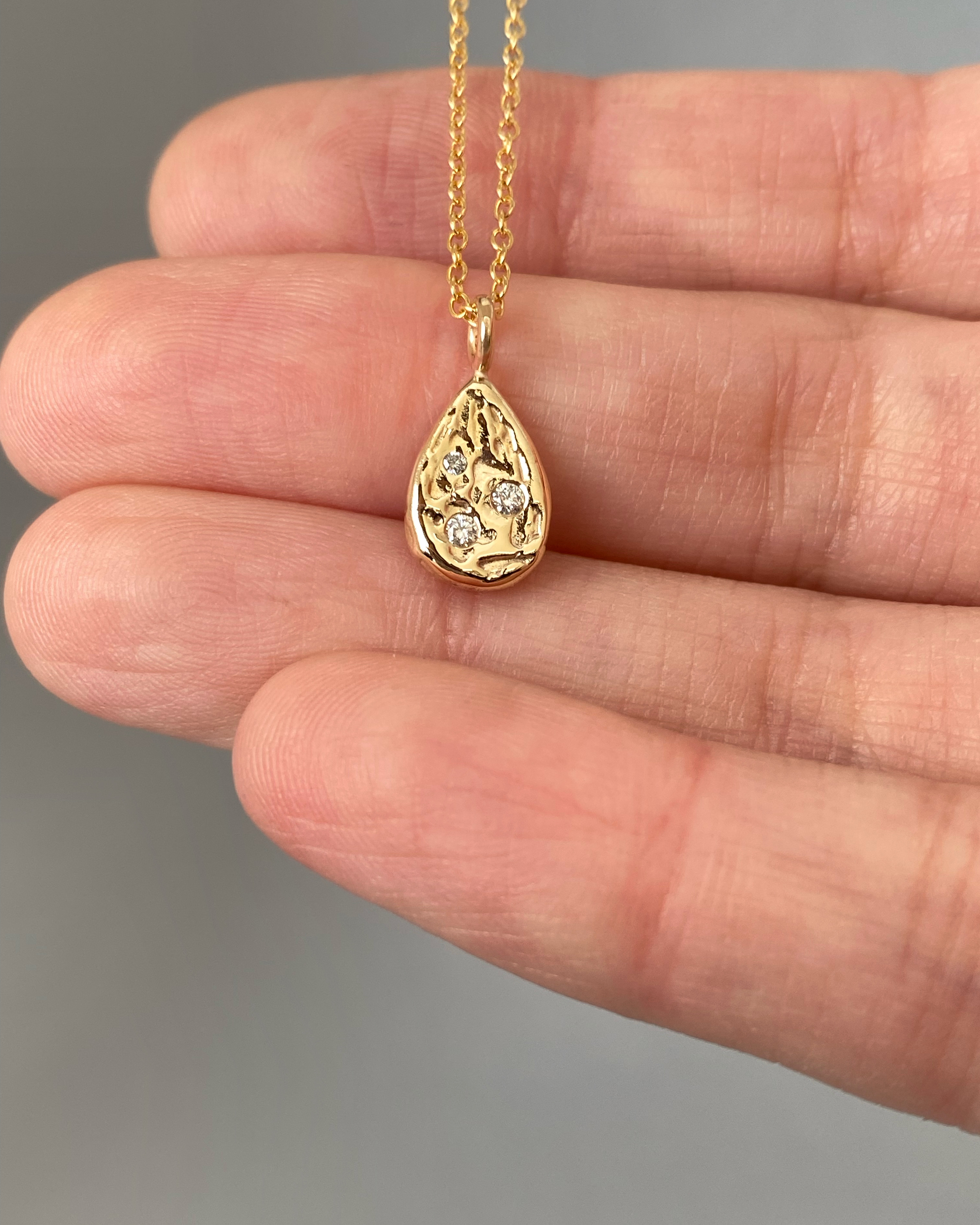 A teardrop-shaped yellow gold pendant with a pitted, organic surface scattered with small flush-set diamonds, held across a palm on a fine gold chain.