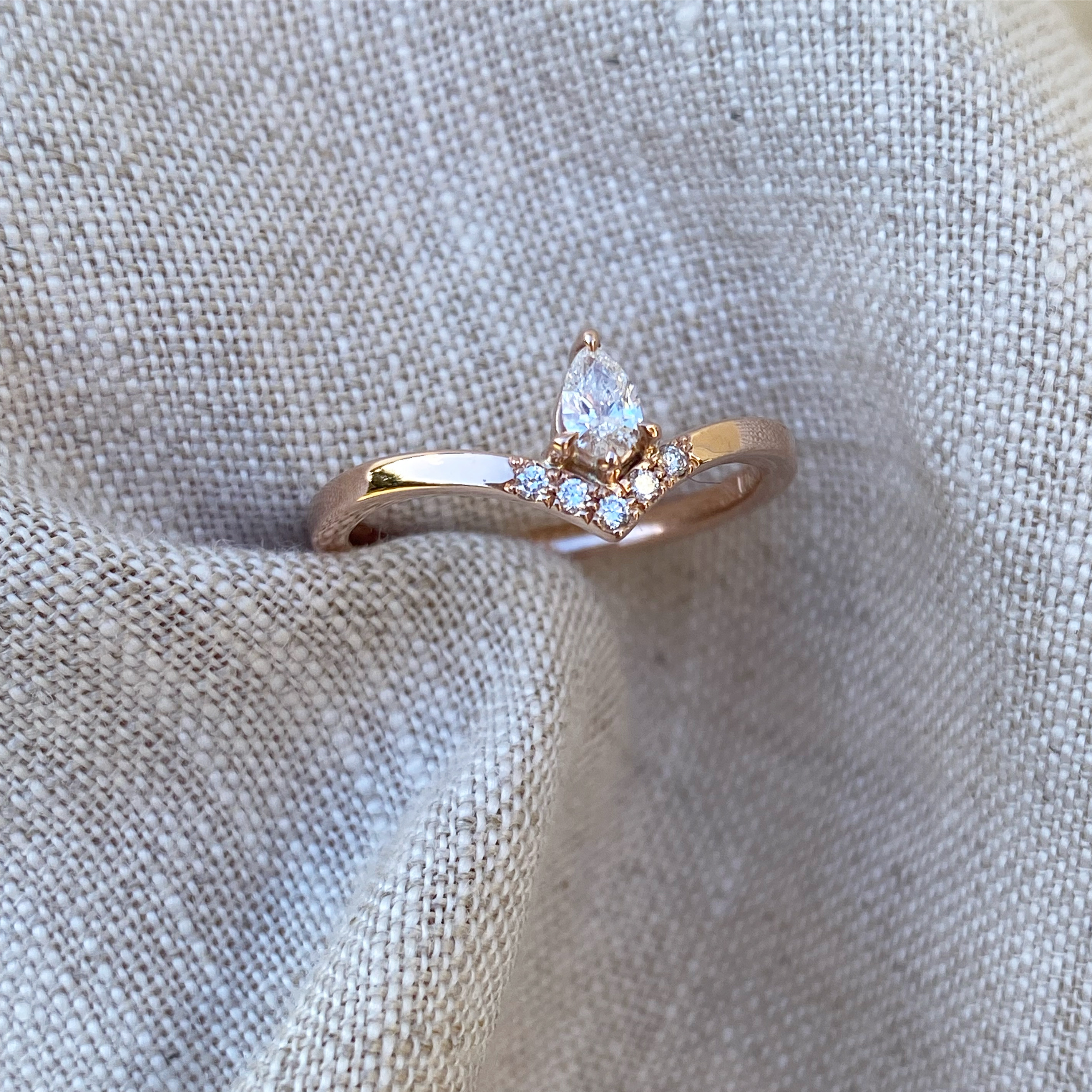 A rose gold ring with a pear-cut diamond above a curved row of smaller round diamonds, set on a band that sweeps up to meet them, on pale linen.