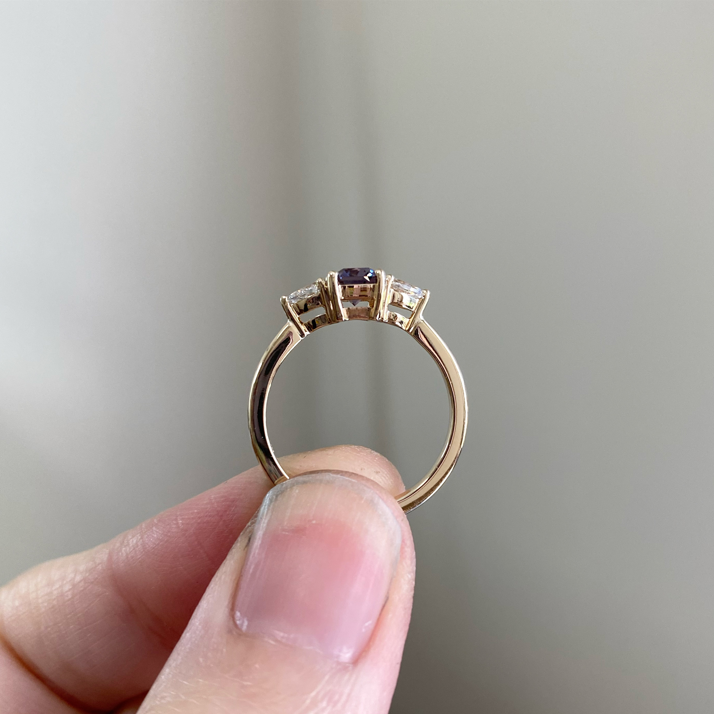 A yellow gold trilogy ring held between finger and thumb, edge on: an oval violet sapphire between two tapered white diamonds.