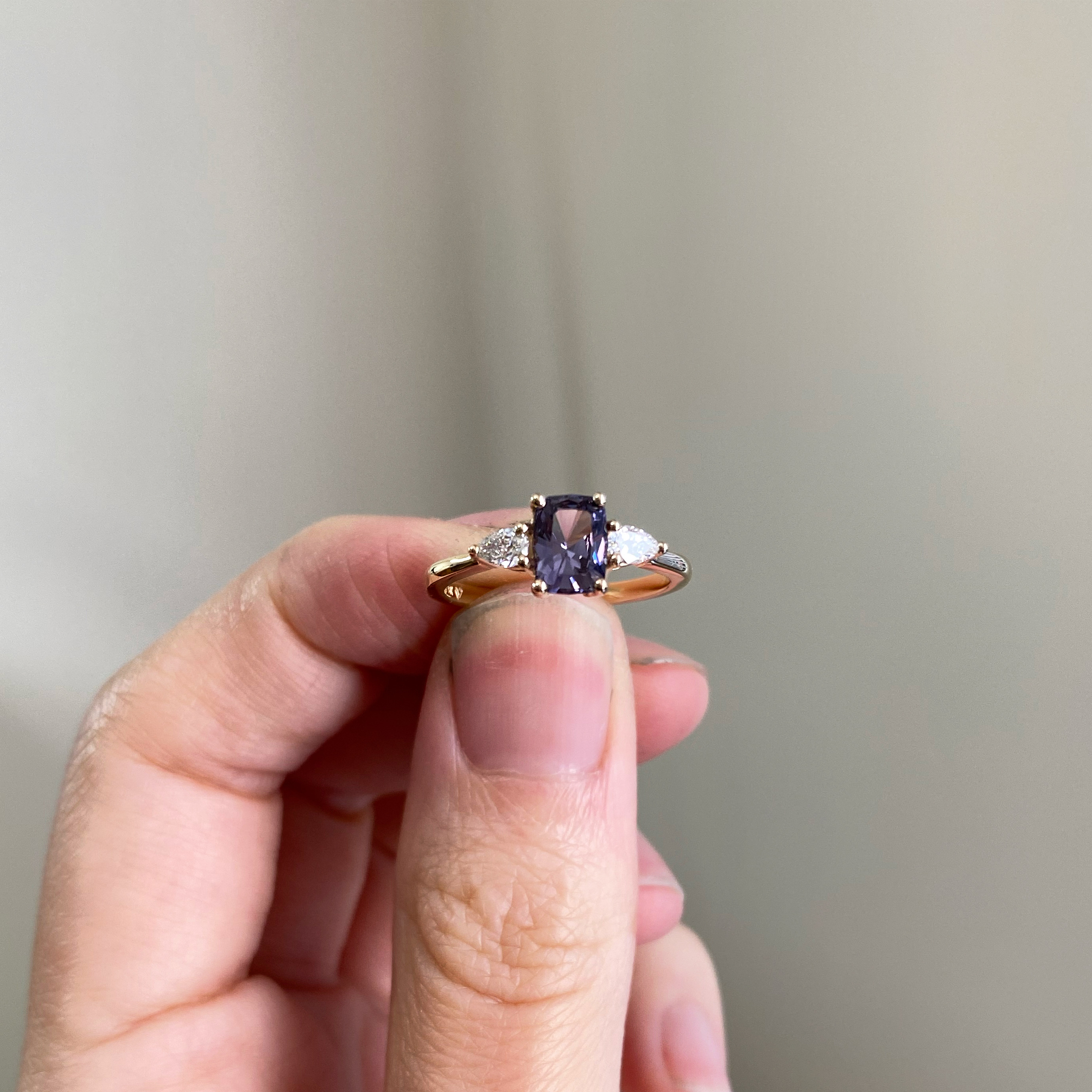 A yellow gold ring held between finger and thumb, set with a cushion-cut violet-grey sapphire flanked by two pear-cut white diamonds.