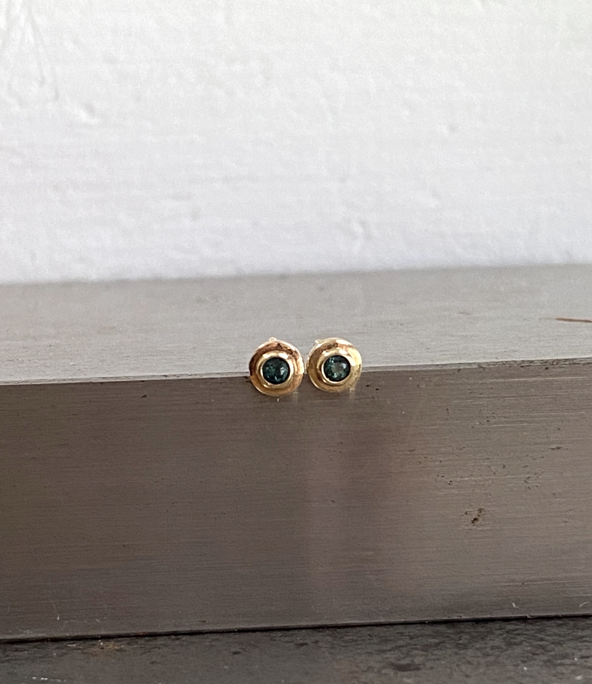 A pair of small yellow gold stud earrings with rough, organic edges, each rubover-set with a dark teal stone, sitting on a grey painted ledge.