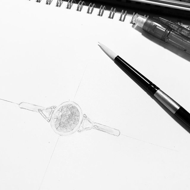 A pencil sketch of a ring on paper: a round faceted stone held by triangular side stones, drawn on a centre line, with a mechanical pencil and a fine brush laid beside it.