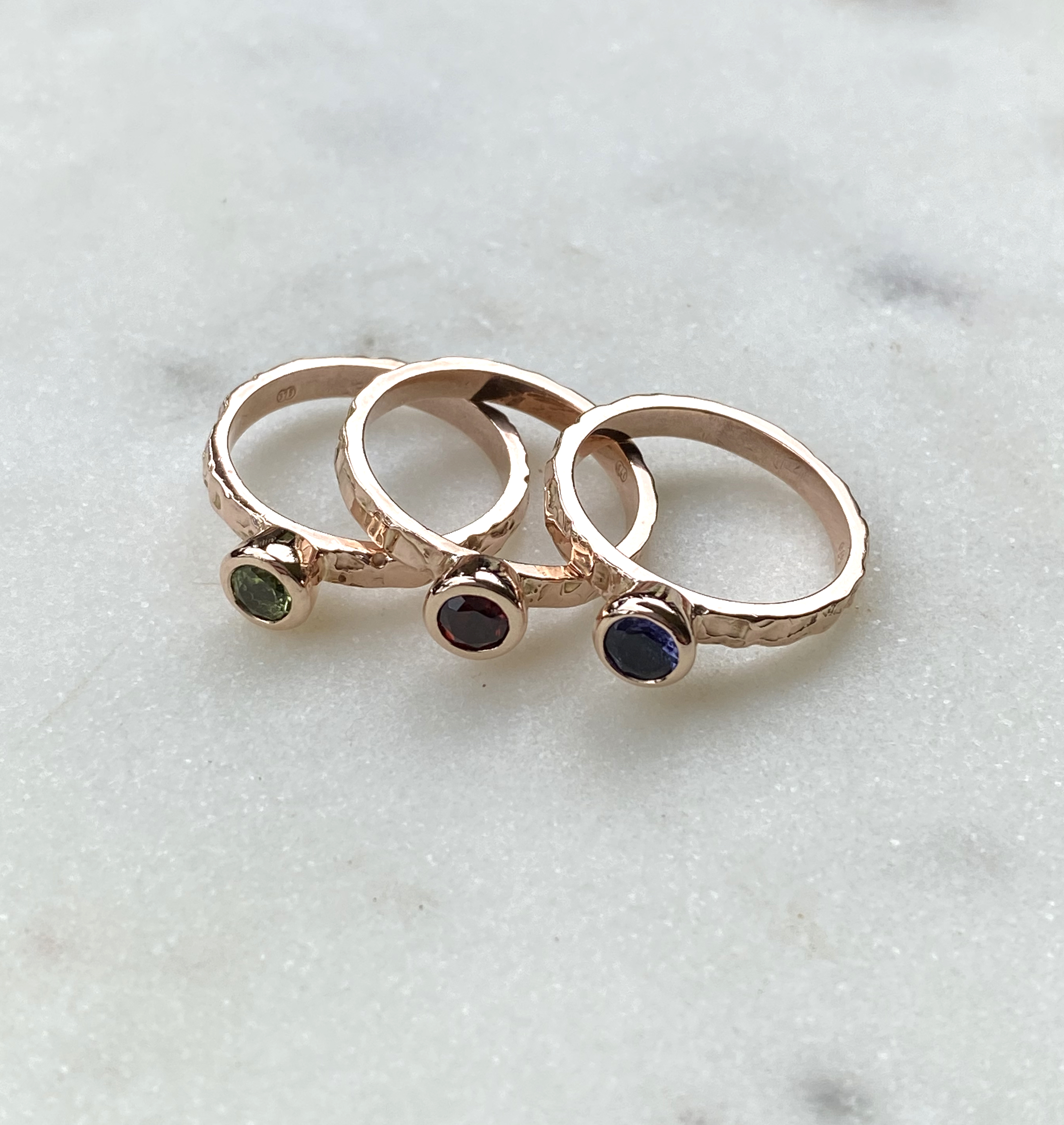 Three hammered rose gold stacking bands side by side, each rubover-set with a single round stone — green, deep red and blue — on white marble.
